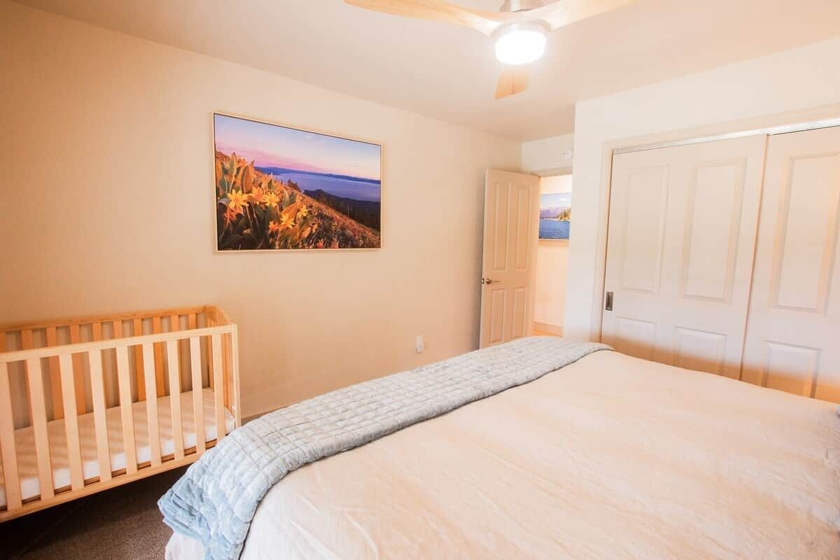 A bedroom with a crib, a bed, a ceiling fan, a door, and a wall-mounted picture.