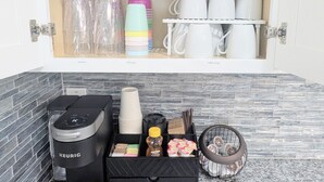 Un meuble de cuisine contenant des verres à vin, des tasses à café et des gobelets pour enfants. En dessous, on trouve une machine à café, une poubelle et un panier avec des ustensiles et des condiments.