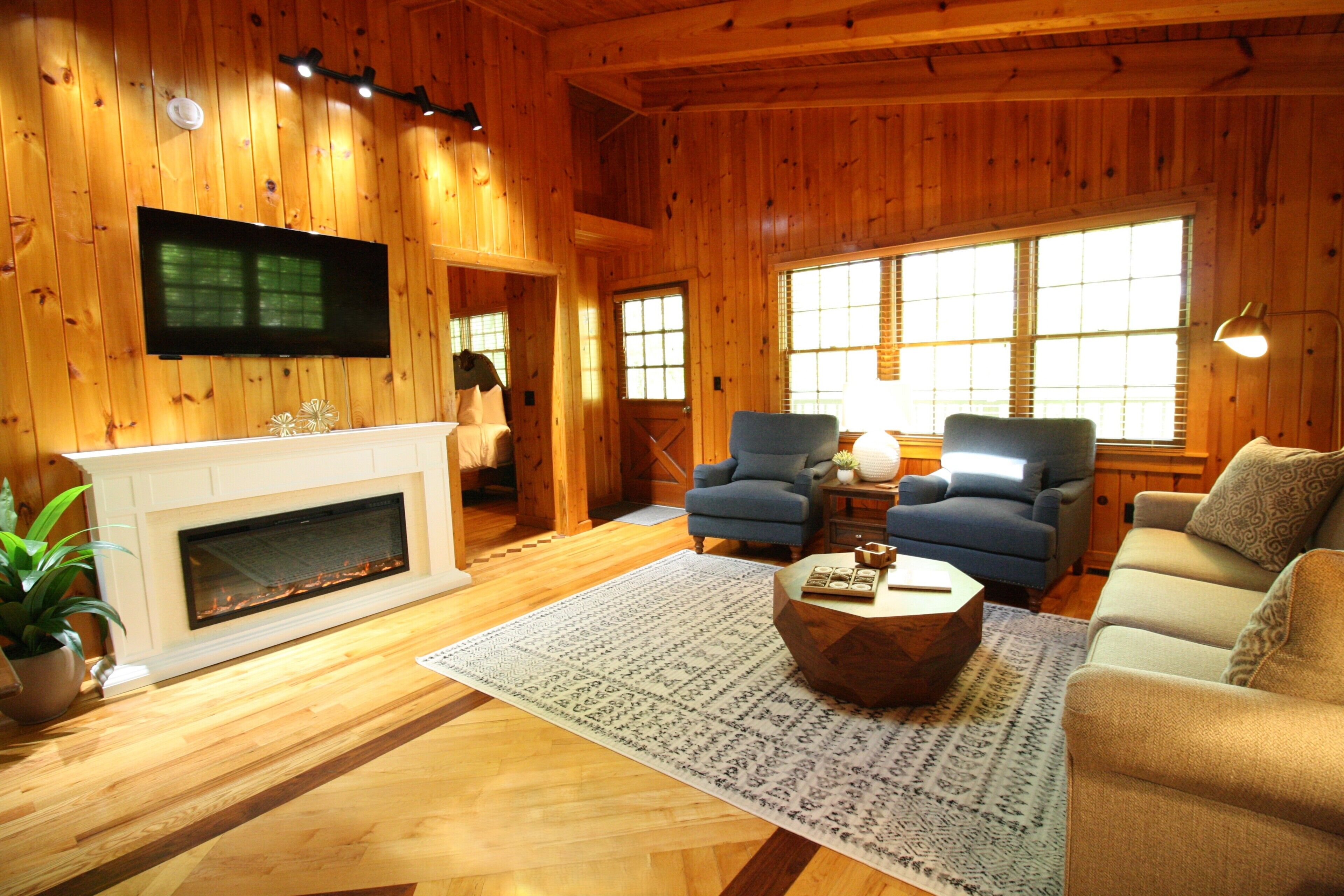 A log cabin interior with a fireplace, two armchairs, a sofa, a television, and a large window.