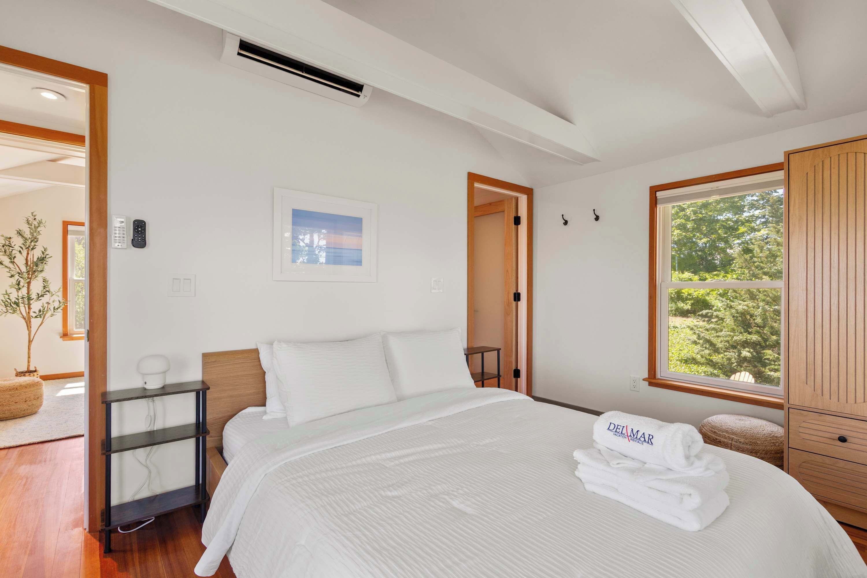 A bedroom with a bed, a bedside table, a wall-mounted light, a window with a view, and a door leading to another room.