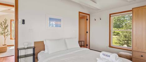 A bedroom with a bed, a bedside table, a wall-mounted light, a window with a view, and a door leading to another room.