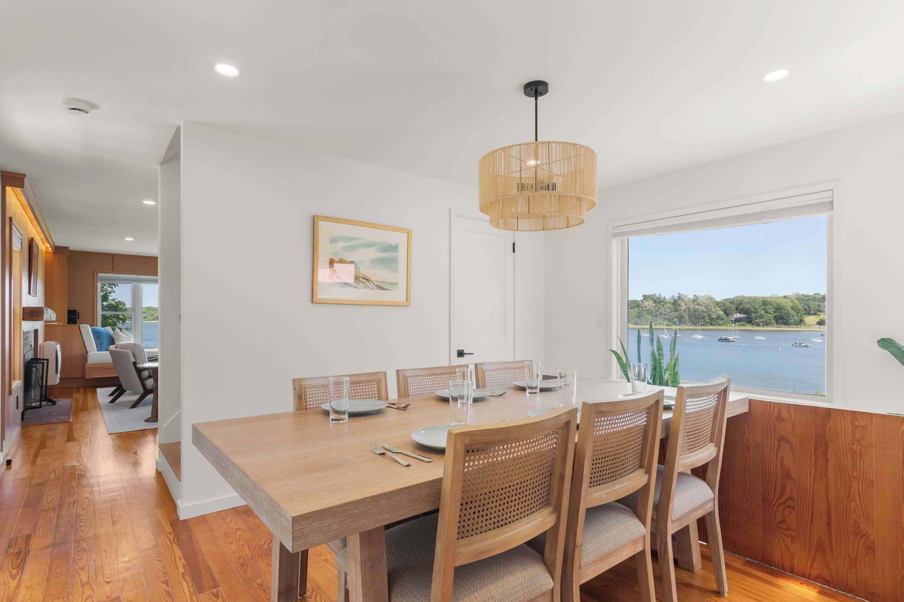Un coin repas avec une table et des chaises en bois, un luminaire suspendu, un tableau encadré au mur et une vue sur l’eau par la fenêtre.