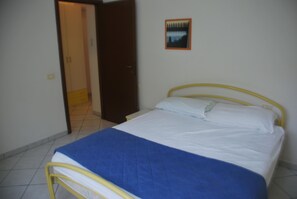 Ein kleines Zimmer mit einem Einzelbett, einem gelben Metallgestell, weißer Bettwäsche und einer blauen Decke.