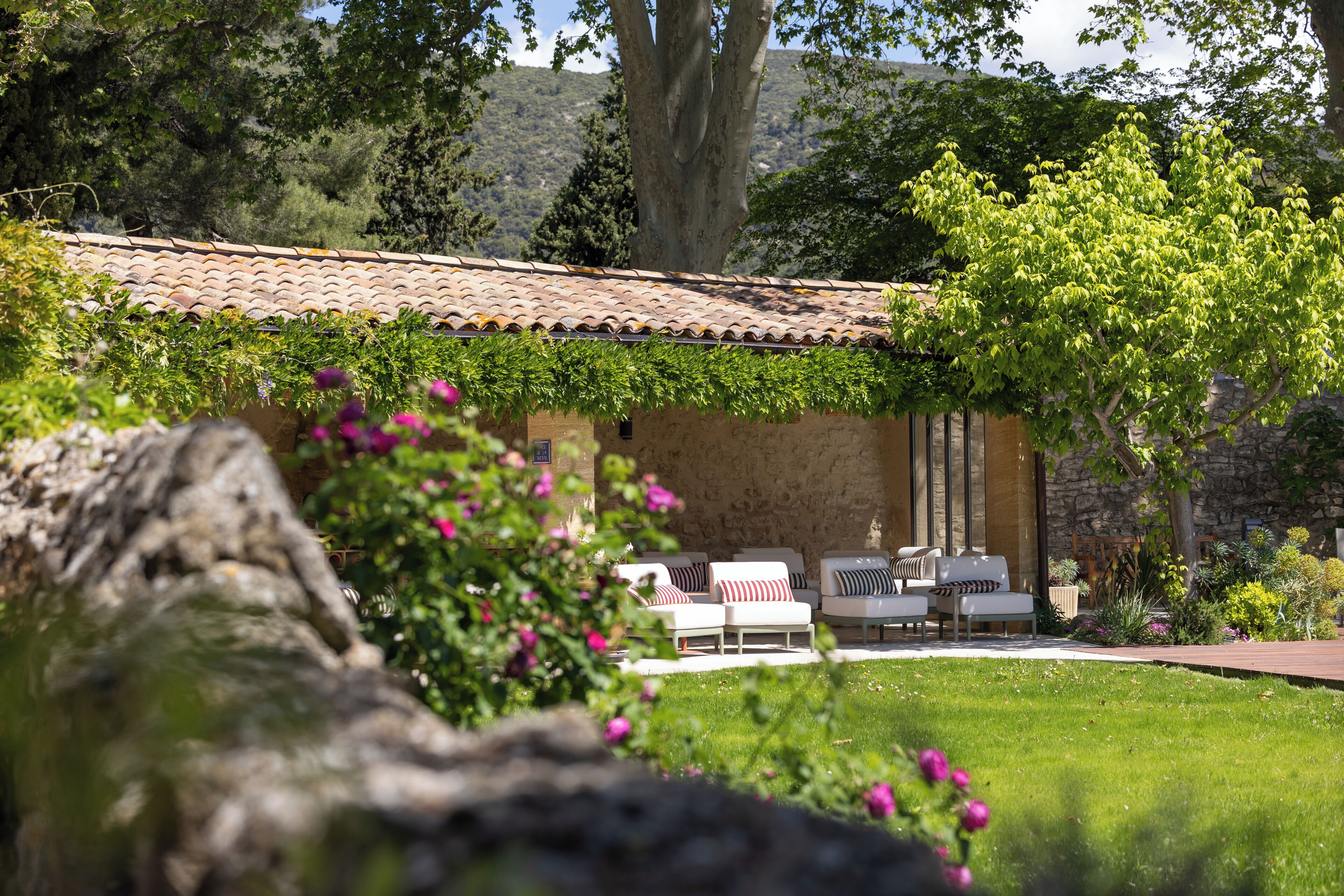 A stone building with a tiled roof, outdoor seating with cushions, and a garden with flowers and trees.