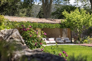 A stone building with a tiled roof, outdoor seating with cushions, and a garden with flowers and trees. - Le Mas de Guilles (Lourmarin)