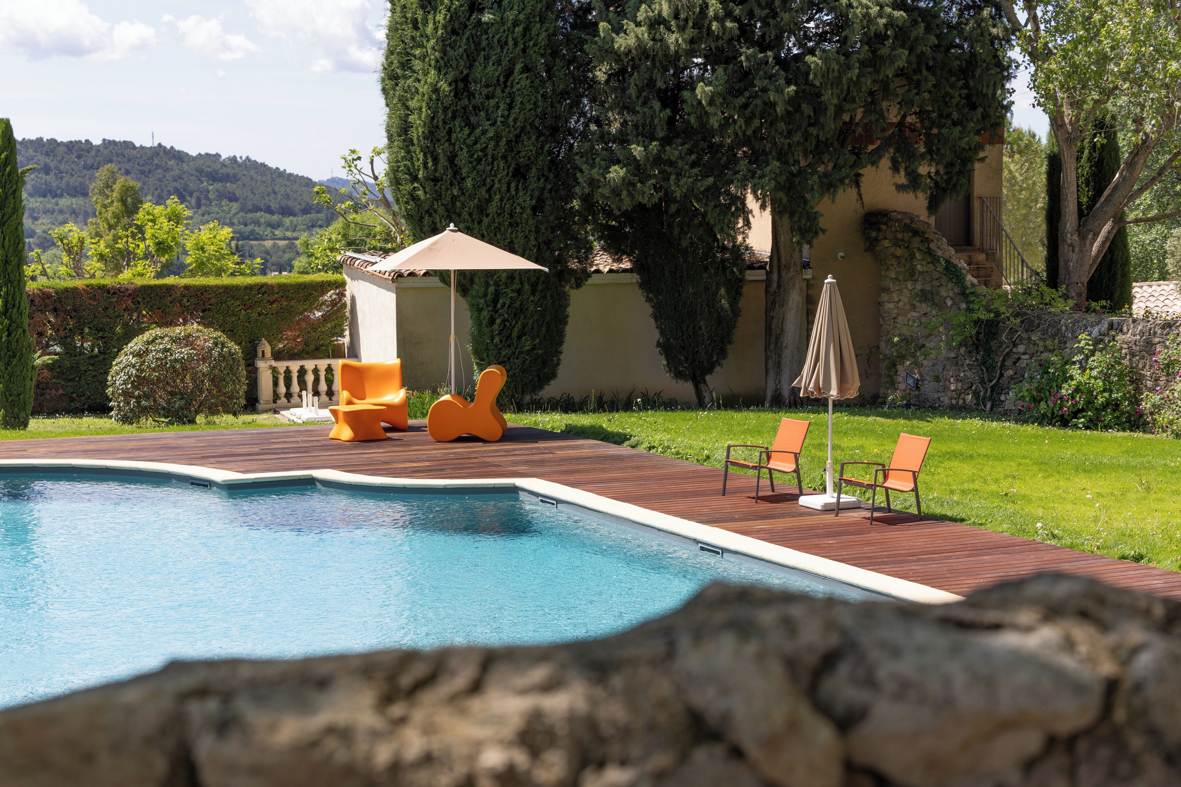 Une piscine entourée d’une terrasse en bois, de chaises longues orange et d’un mur en pierre.