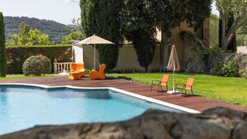 Une piscine entourée d’une terrasse en bois, de chaises longues orange et d’un mur en pierre.