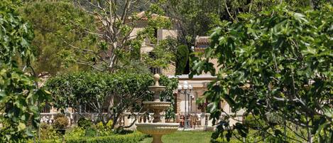 A garden with a fountain, neatly trimmed hedges, and a building with a tiled roof in the background.
