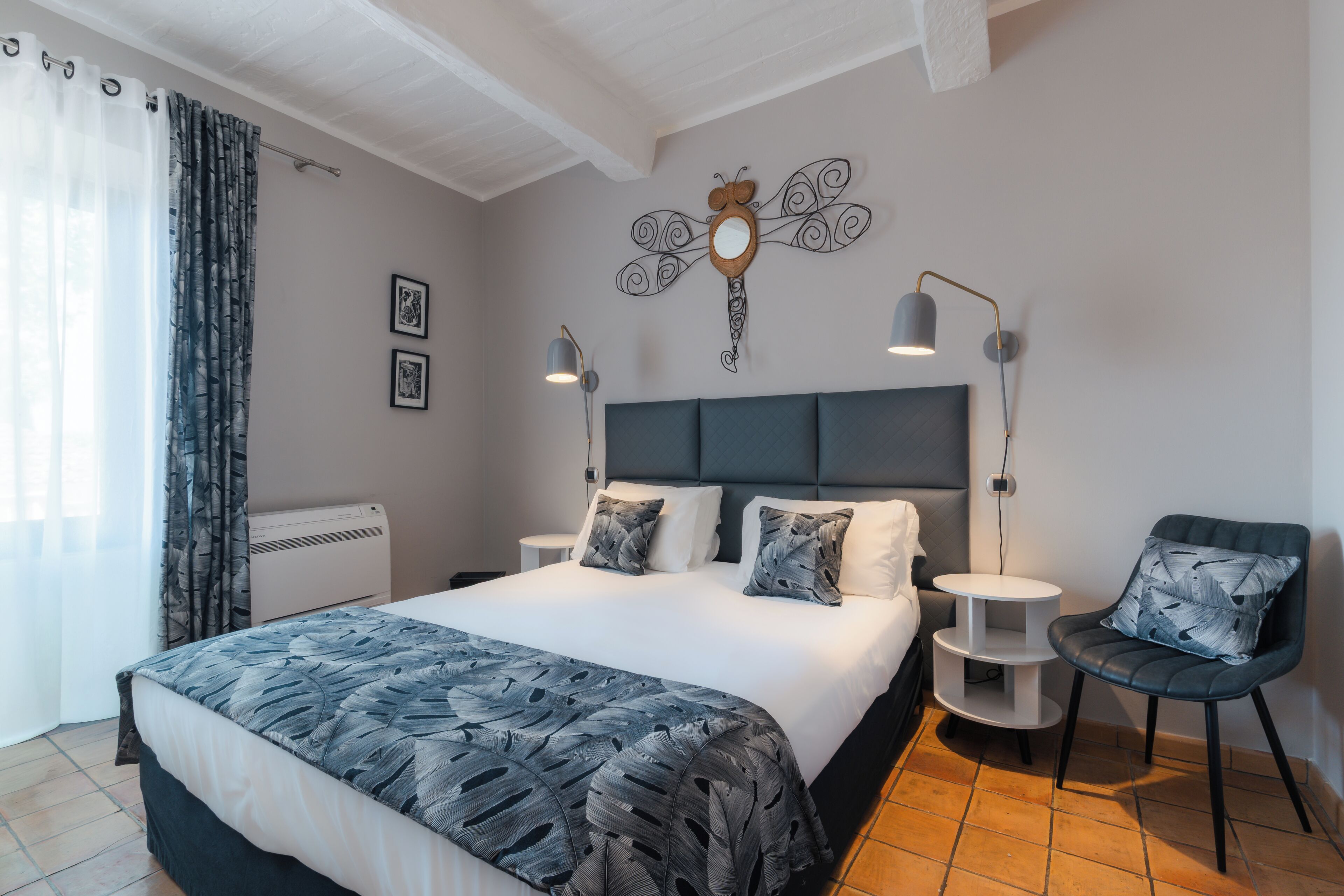 A modern hotel room with a large bed, two bedside tables, a chair, a decorative wall piece, and a window with curtains.