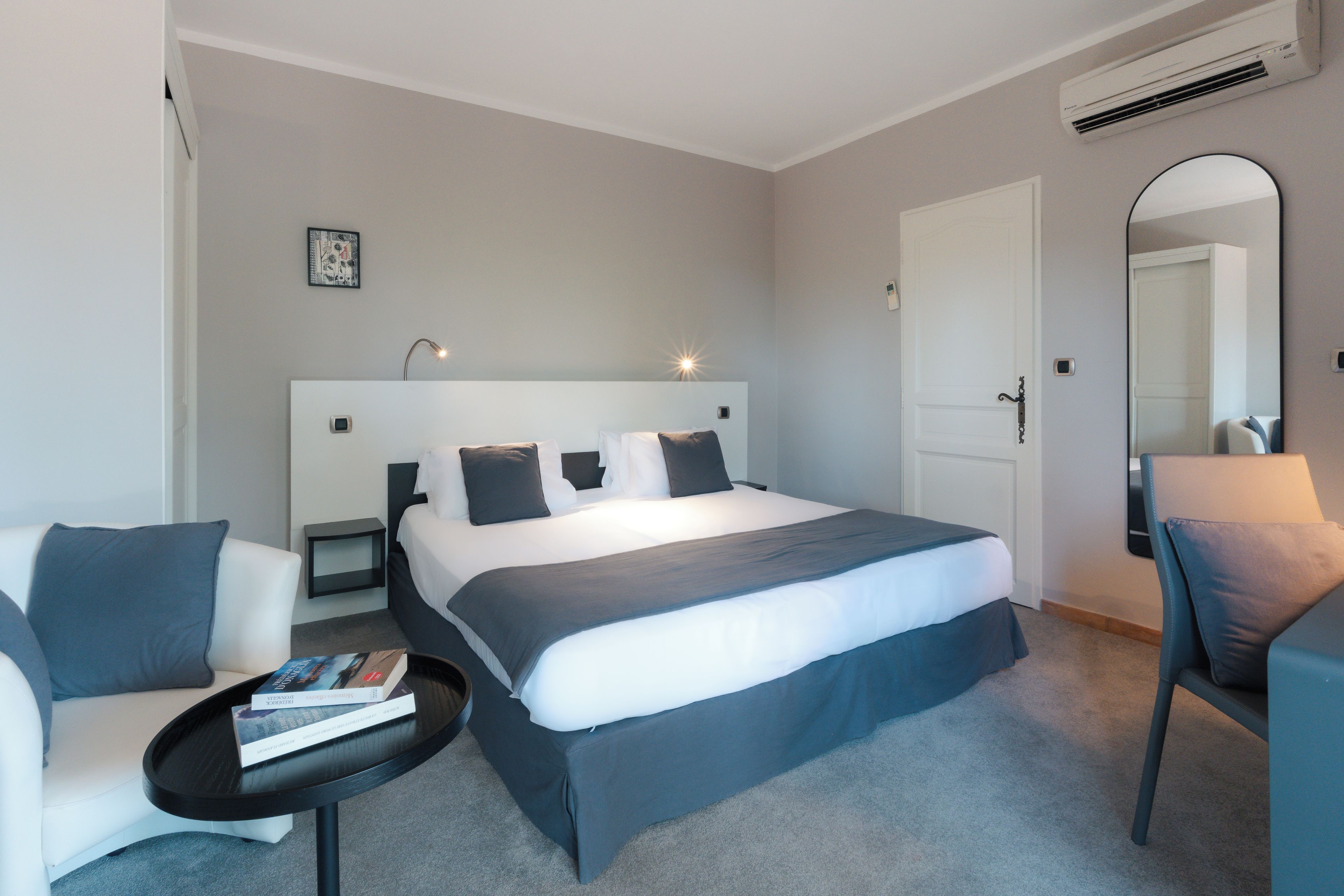 Une chambre d’hôtel moderne avec un grand lit, des tables de chevet, une chaise, une petite table avec des livres et un miroir.