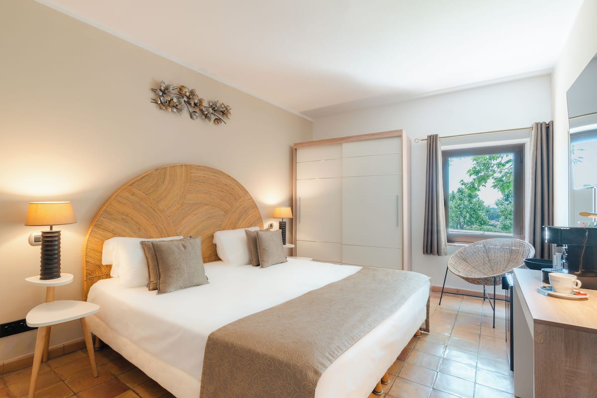 Une chambre d’hôtel avec un grand lit, des lampes de chevet, un bureau avec une chaise et une vue sur la verdure par la fenêtre.