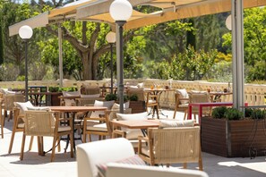 An outdoor patio with wooden tables and chairs, a covered seating area, and a view of trees and greenery. - Le Mas de Guilles (Lourmarin)