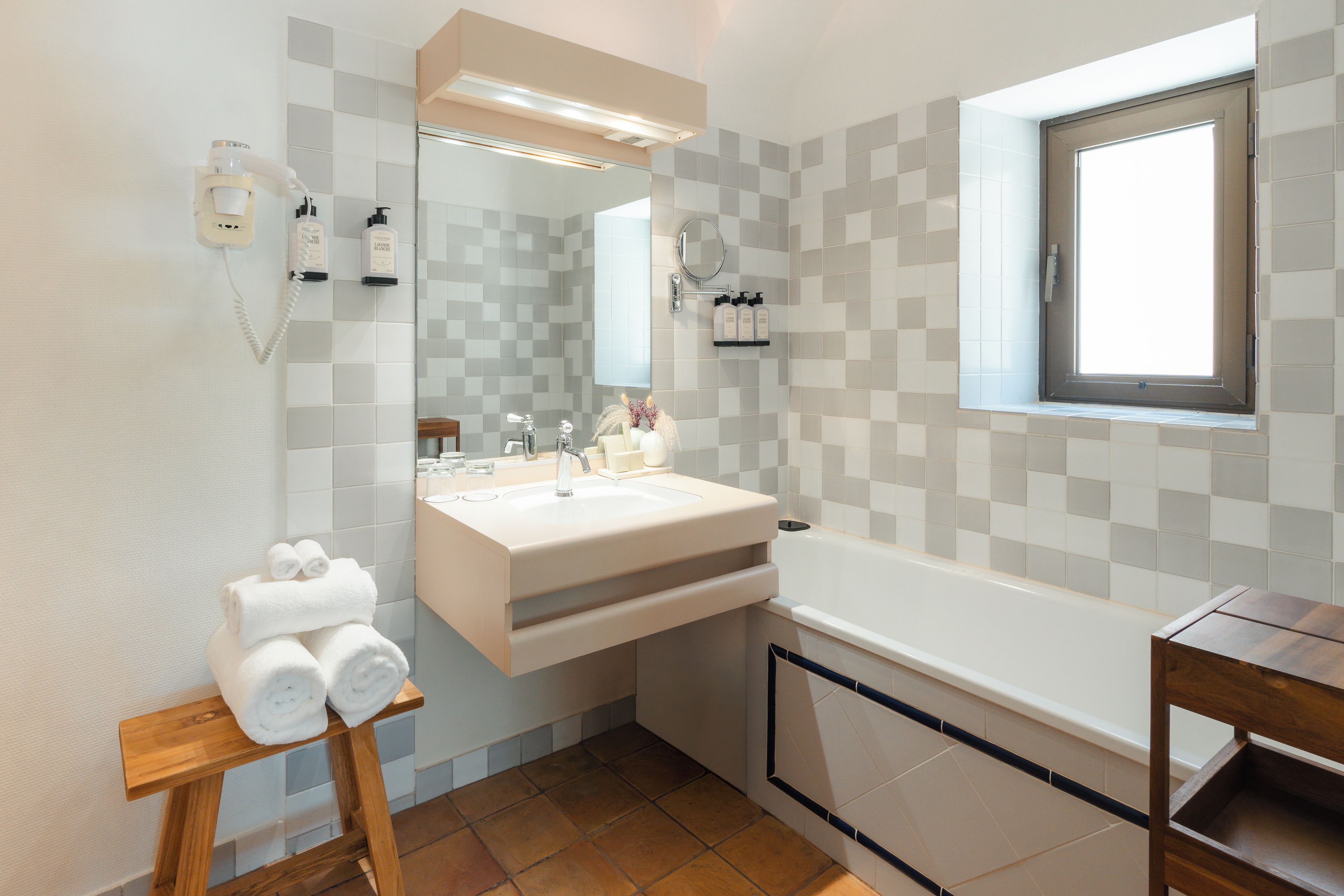 A modern bathroom with a large mirror, a white sink, a bathtub, and a wooden stool with towels.