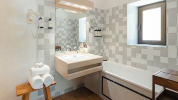 A modern bathroom with a large mirror, a white sink, a bathtub, and a wooden stool with towels.