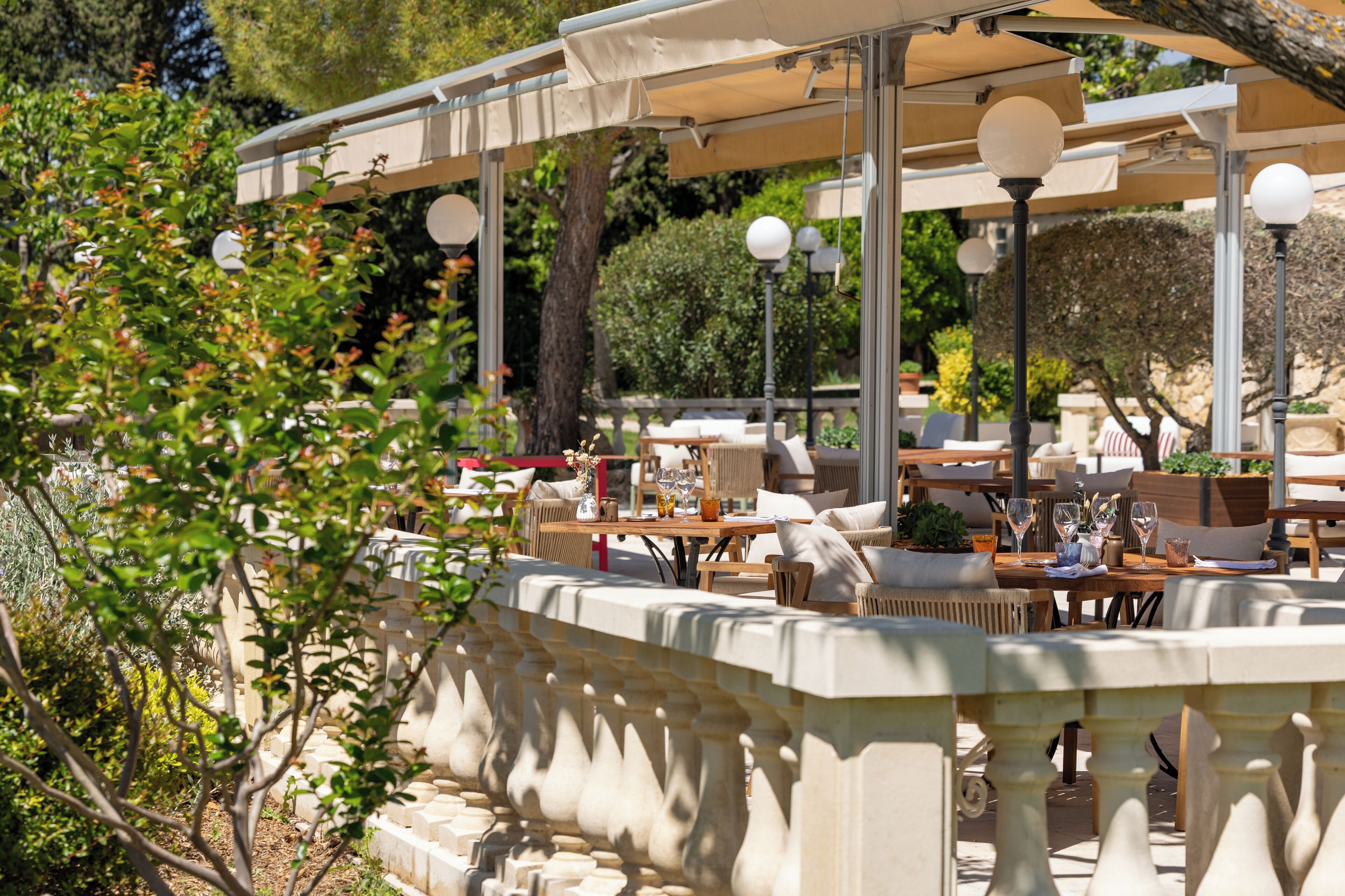 An outdoor café with tables and chairs, surrounded by greenery and a stone balustrade.