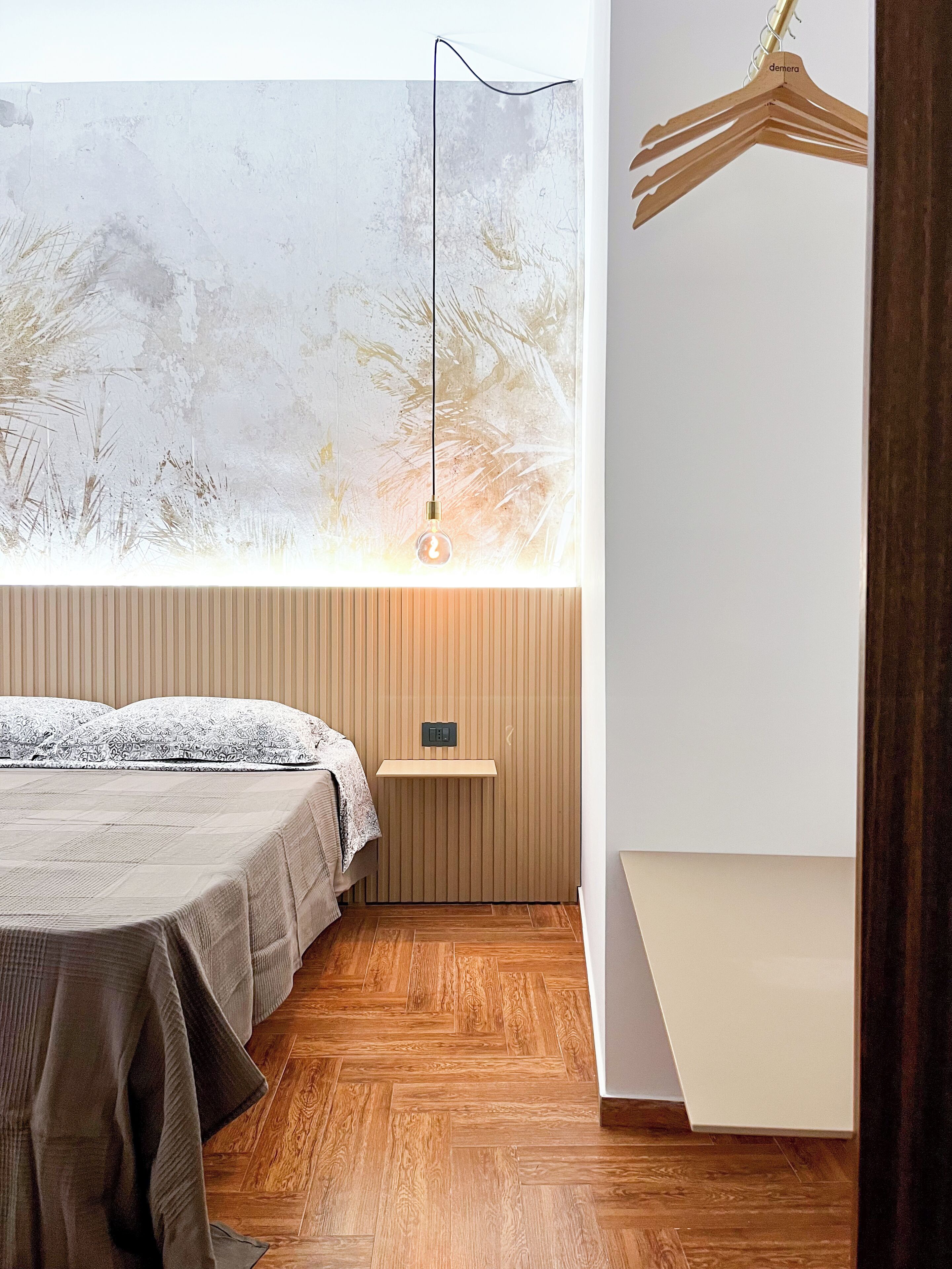 A bedroom with a bed, a hanging light, a wooden shelf, and a view of trees through a window.