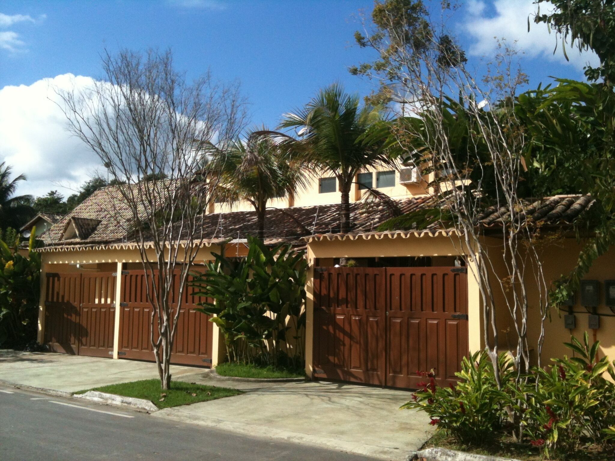 A residential house with two garages, a tiled roof, and a front yard with palm trees and shrubs.