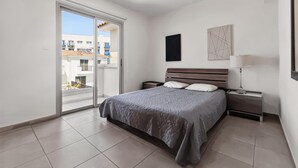 Ein Schlafzimmer mit Bett, Nachttisch, Lampe, einer Schiebetür zum Balkon und Blick auf Gebäude.
