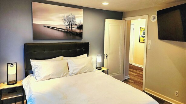 A hotel room with a bed, a nightstand, a wall-mounted TV, a door, and a painting above the bed. - 513Company - Theater +Location (Cincinnati)