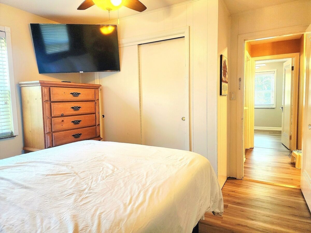 Ein Schlafzimmer mit einem Bett, einer Kommode, einem an der Wand montierten Fernseher und einem Flur, der in ein weiteres Zimmer führt.