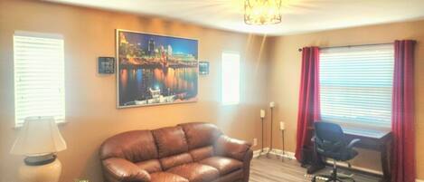 A room with a brown leather sofa, a desk with a chair, a cityscape painting, and a chandelier.