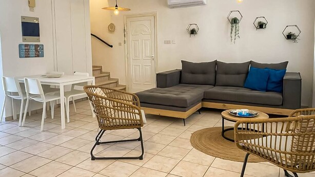 Un salon moderne avec un canapé d’angle gris, deux fauteuils en osier, une table à manger avec des chaises et un escalier menant à un autre niveau.