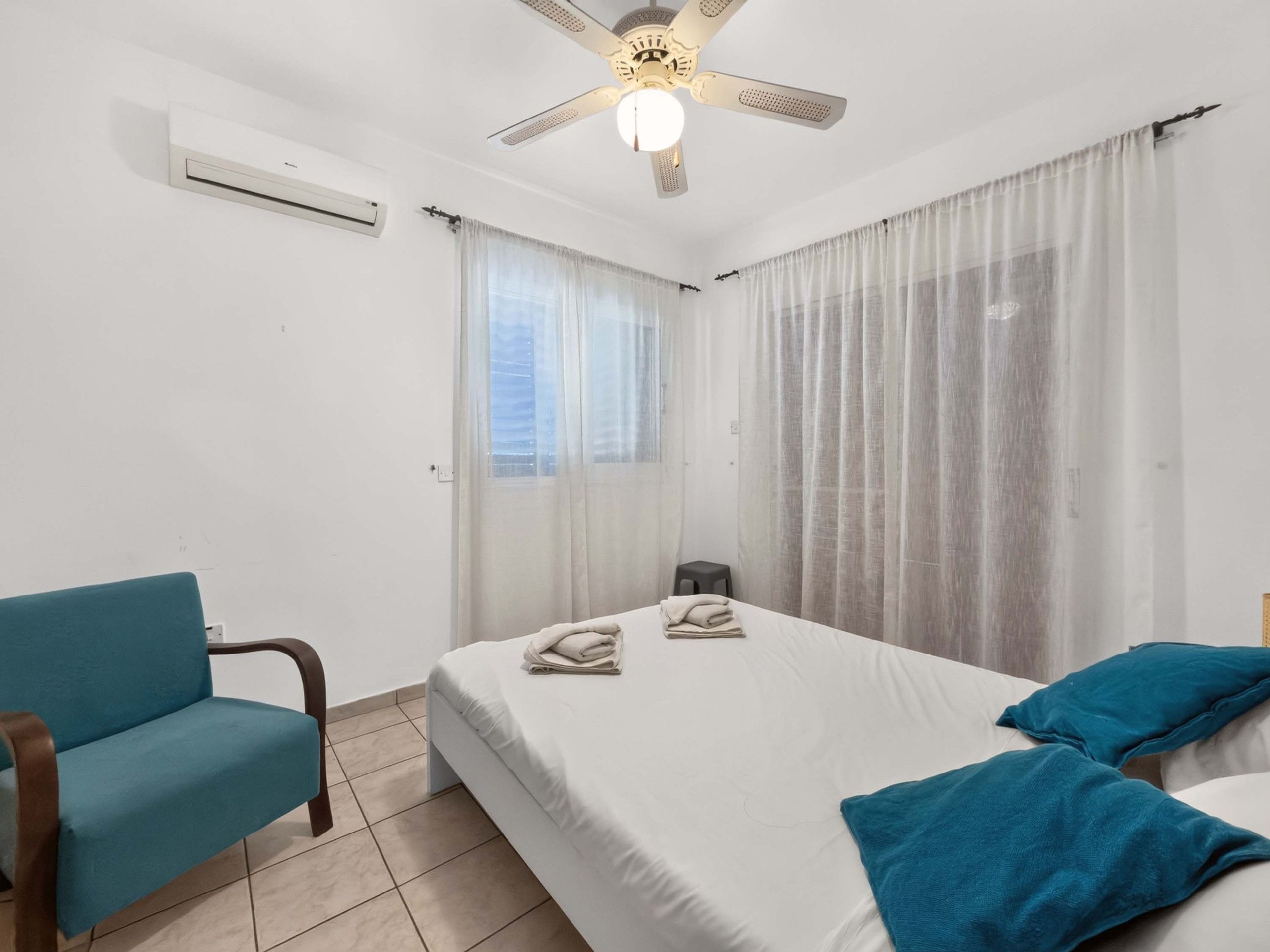 Un dormitorio con cama, una silla, un ventilador de techo y una ventana con cortinas.