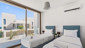 A modern hotel room with two beds, a large window with a view of buildings, an air conditioning unit, and a ceiling light. - Azure Sama Villa – Jacuzzi / Heated Pool (Protaras)