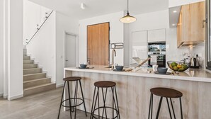 A modern kitchen with a central island, bar stools, and a staircase leading to another level. - Azure Sama Villa – Jacuzzi / Heated Pool (Protaras)