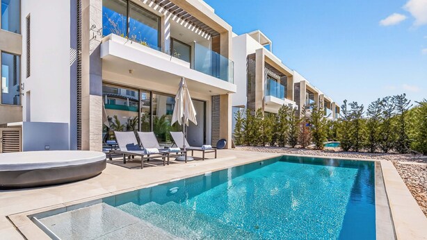 A modern poolside area with a clear pool, lounge chairs, and a building with balconies. - Azure Sama Villa – Jacuzzi / Heated Pool (Protaras)