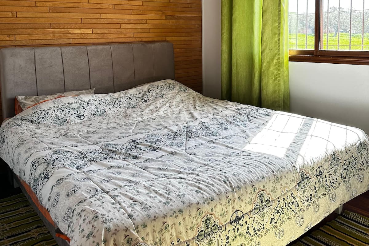 A bedroom with a wooden wall, a grey headboard, a patterned bedspread, a window with green curtains, and an air conditioning unit.