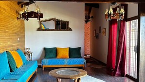 Un salon chaleureux avec un plafond en bois, un canapé bleu agrémenté de coussins colorés, une table basse ronde en bois et un mur en briques en arrière-plan.