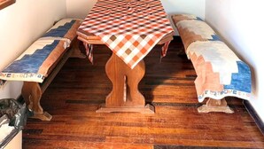 Una habitación de esquina con una mesa y bancos de madera, techo de madera y una estantería con cerámica.