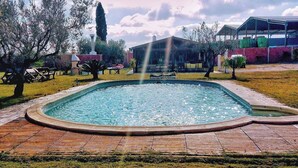 Una piscina con fuente, rodeada de césped y árboles, con un edificio al fondo.