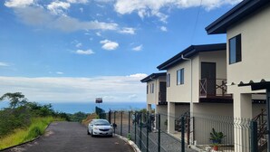 A modern two-story house with a car parked in front, a fenced garden, and a clear view of the ocean. - Casa Pura Vista (Playa Hermosa)