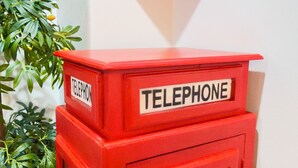 Une cabine téléphonique rouge, portant une étiquette, est posée sur une table en bois devant un mur blanc.
