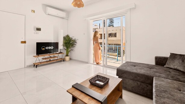 Een moderne woonkamer met een flatscreen televisie, een houten salontafel, een grijze bank en een grote schuifpui die leidt naar een buitenbalkon.