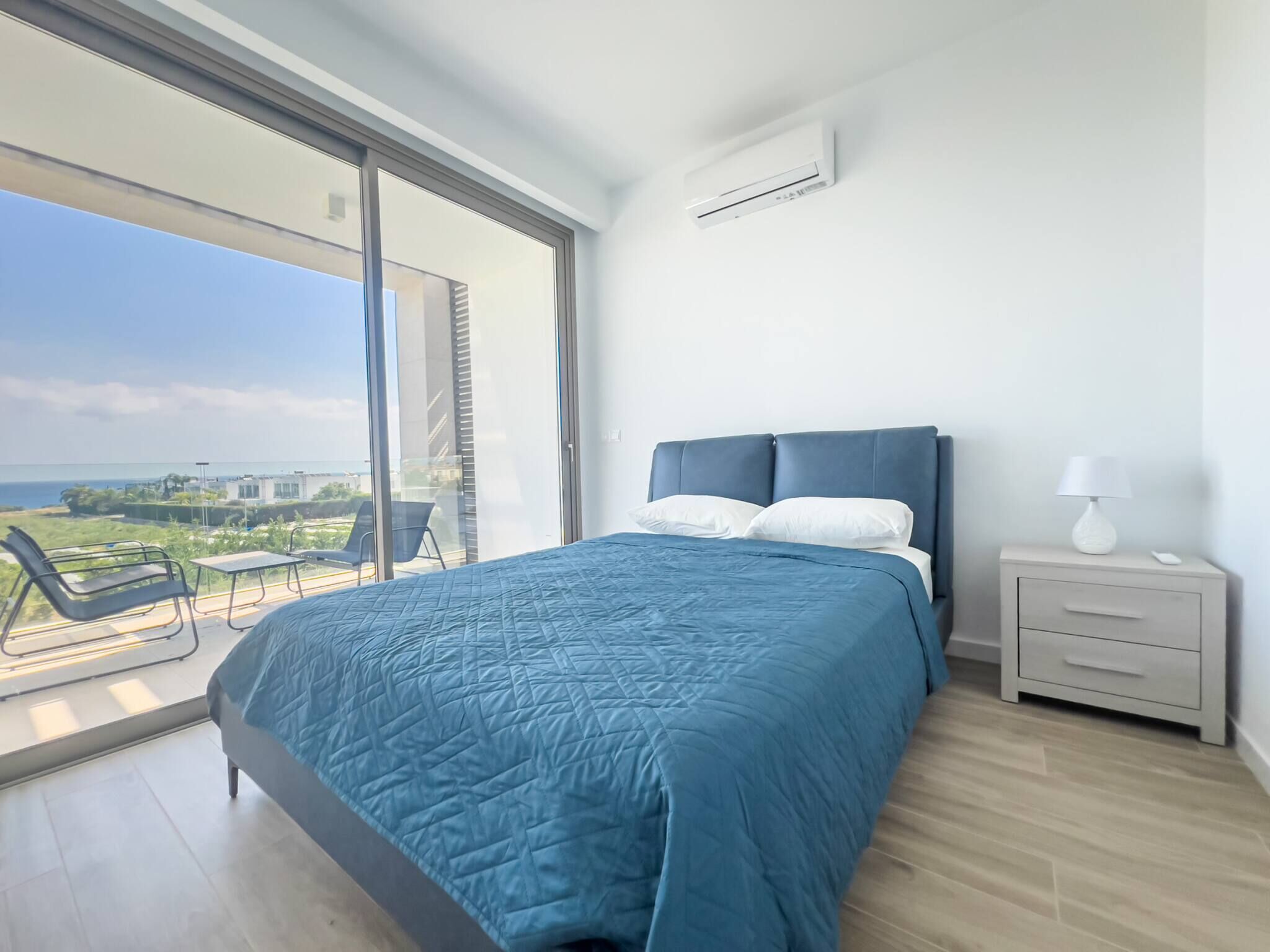 Un dormitorio moderno con una cama grande, una mesita de noche, una lámpara y vistas al mar a través de un ventanal corredero.