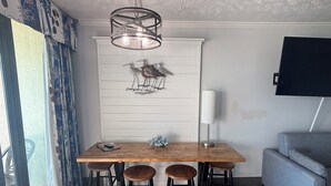 Un coin repas avec une table en bois, quatre tabourets, un luminaire suspendu et un téléviseur fixé au mur.