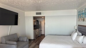 Une chambre d’hôtel moderne avec un canapé gris, un grand lit avec une literie blanche, un téléviseur à écran plat fixé au mur et un coin cuisine.