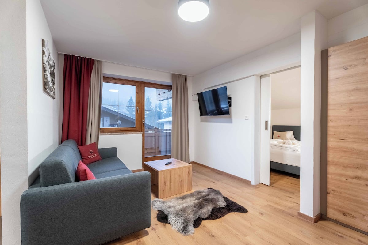 A compact living space with a sofa, a small wooden table, a TV mounted on the wall, and a bed in the adjacent room.