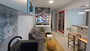 A compact living space with a grey sofa, a small white table, a patterned ottoman, a TV mounted on the wall, and a red-framed glass door leading to a kitchen area. - Bagnol Chill - Your cosy cocoon 30 min from Avignon (4 pers.) (Bagnols-sur-Cèze)