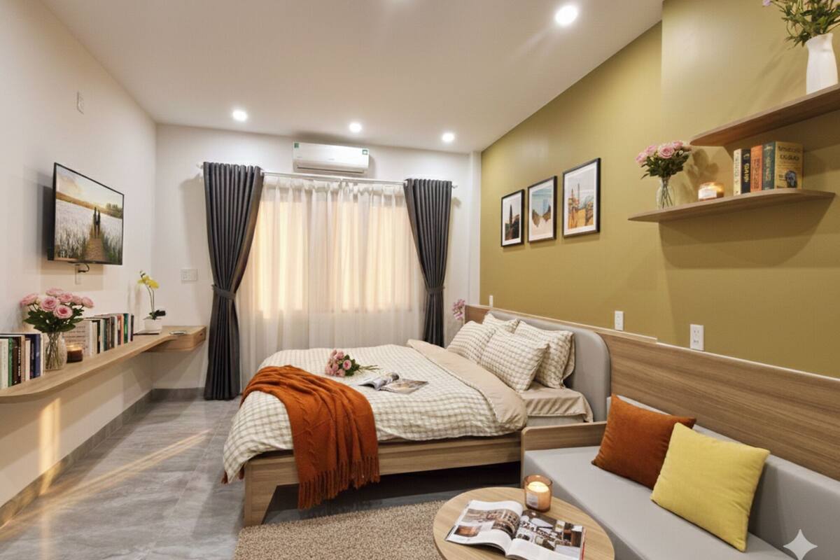 Un dormitorio moderno con cama, sofá, mesita de noche con revista, estante de pared con libros, ventana con cortinas y equipo de aire acondicionado.