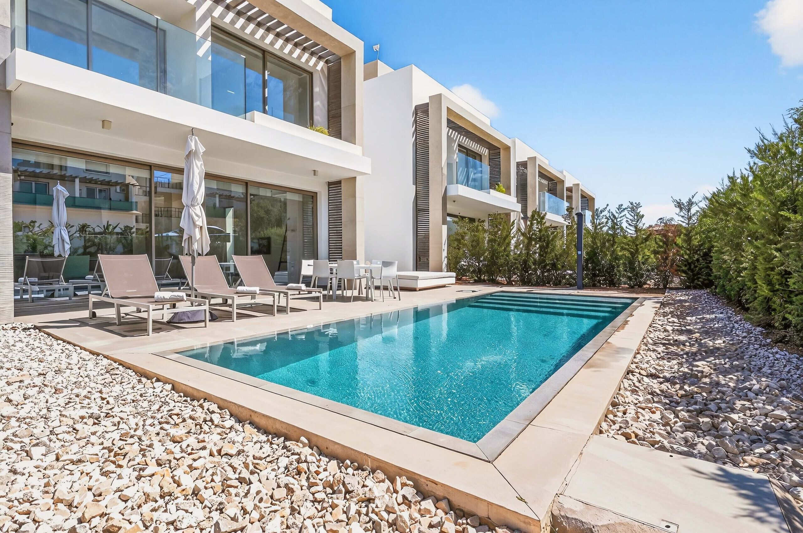 A modern pool area with lounge chairs and umbrellas, surrounded by a pebble-lined deck and a building with large windows and balconies.