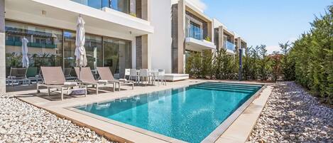 A modern pool area with lounge chairs and umbrellas, surrounded by a pebble-lined deck and a building with large windows and balconies.