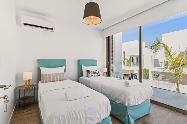 Une chambre d’hôtel avec deux lits, une table de chevet avec une lampe, une grande fenêtre donnant sur des immeubles et des palmiers, et un appareil de climatisation fixé au mur.