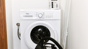 Lavadora blanca compacta con panel de control y manguera, ubicada en un pequeño espacio de lavandería con piso de baldosas.
