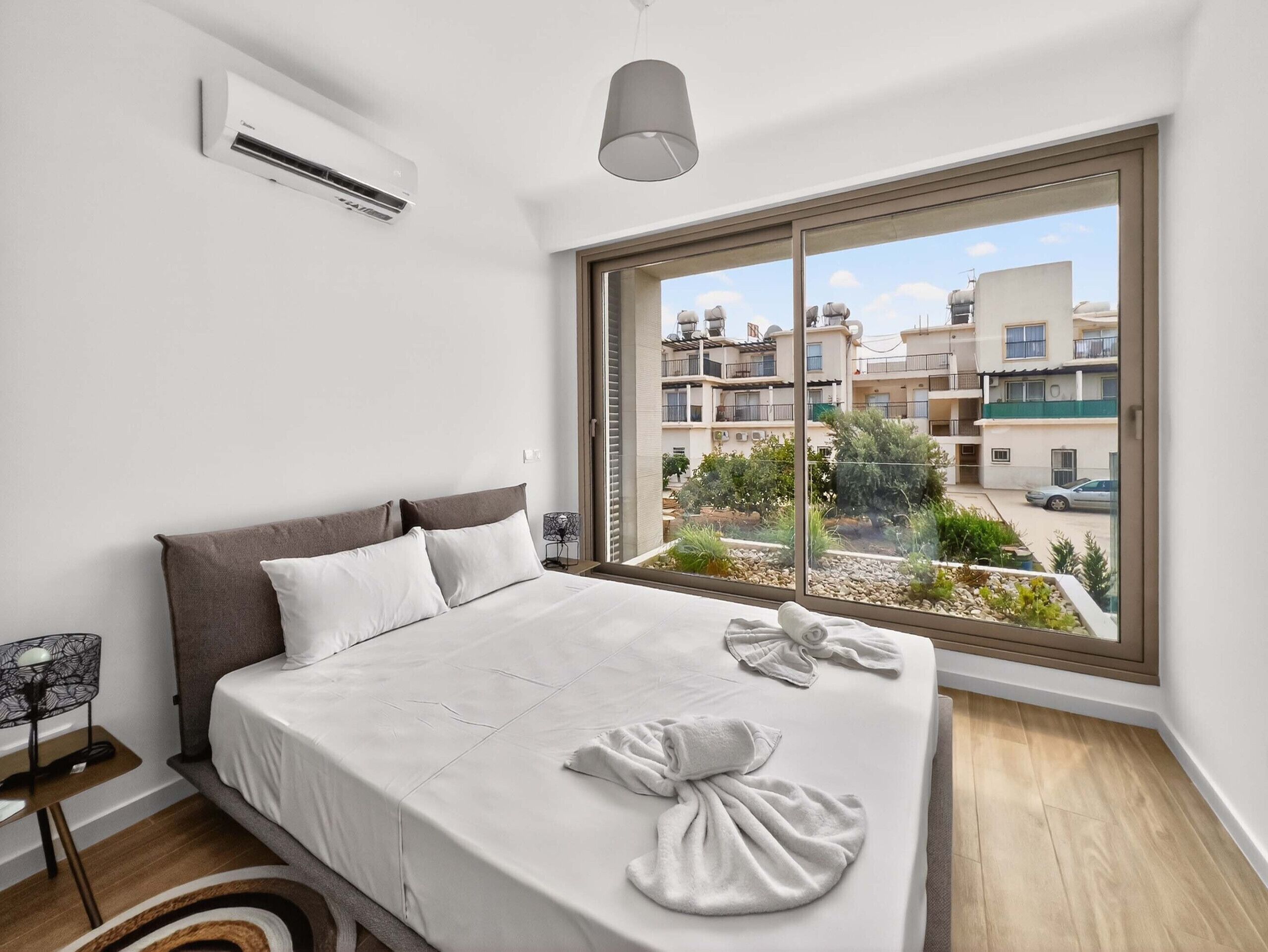 Ein Schlafzimmer mit großem Fenster, einem Bett mit weißer Bettwäsche, einem Nachttisch und einer Klimaanlage.
