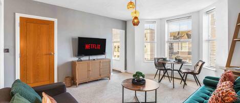 A modern living room with a sofa, a coffee table, and a TV displaying 'NETFLIX'. There is a dining area with a table and chairs, and a wooden cabinet.