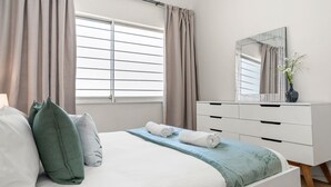 A neatly made bed with white and green pillows, a light blue bedspread, and a white dresser with a mirror and vase. - Art Haus Sea Point (Cape Town)